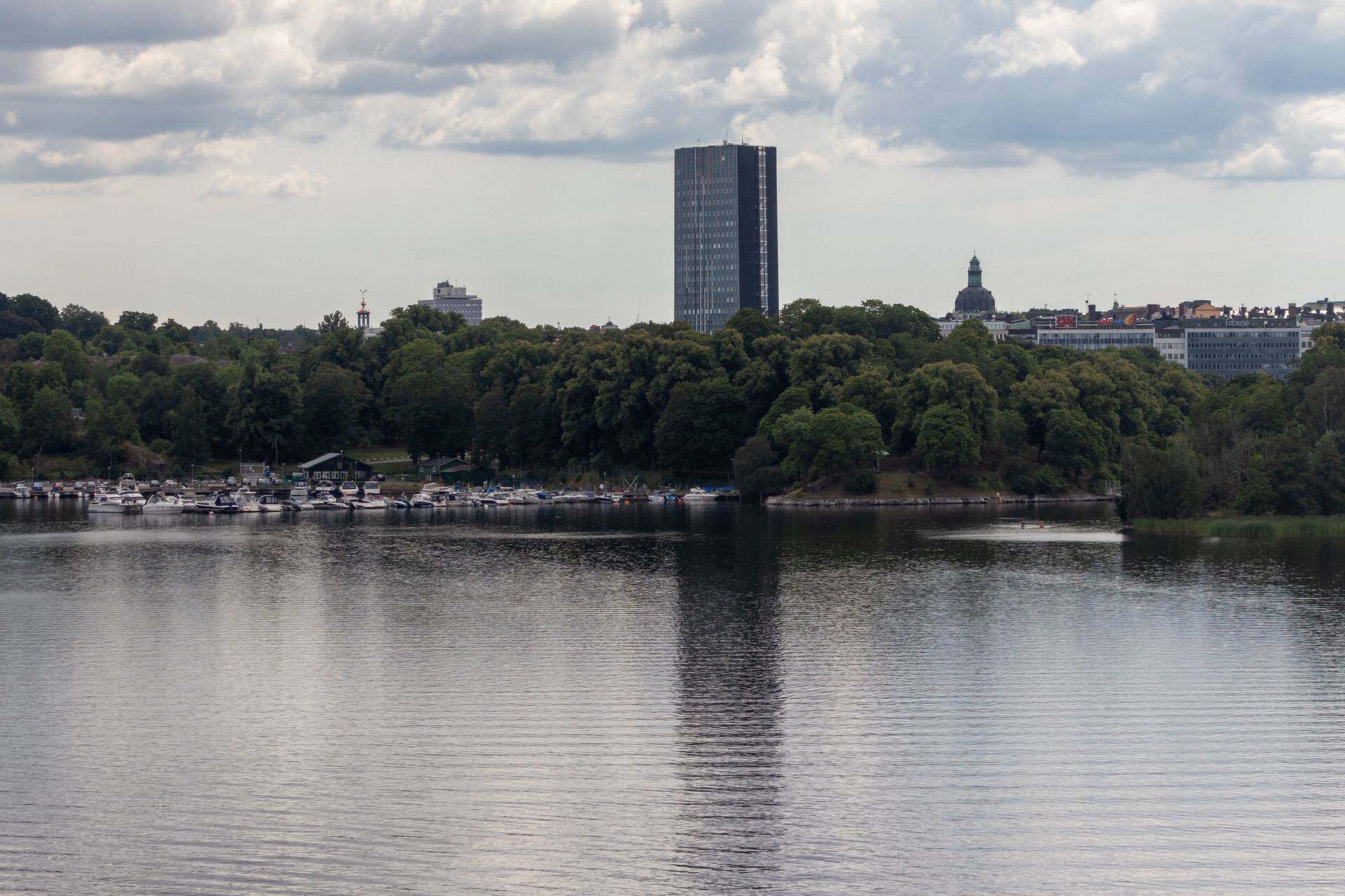 En bild av en stadssilhuett med höga byggnader och en sjö i förgrunden, under en molnig himmel, som visar en urban miljö med naturinslag.
